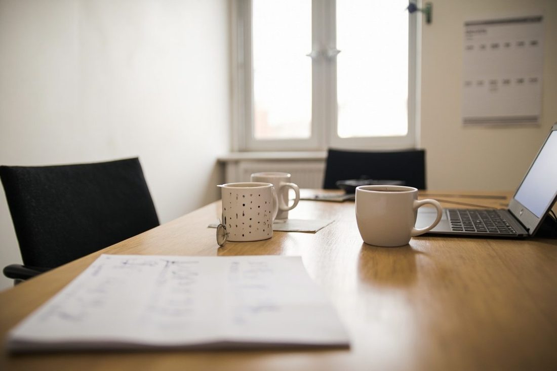 A quiet office meeting room with two empty chairs at a wooden table, mugs of tea, an open laptop and a printed sheet of notes, lit by daylight from a window