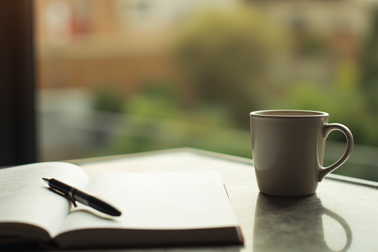 Open notebook and pen beside a mug of tea on a kitchen counter overlooking a back garden in soft morning daylight