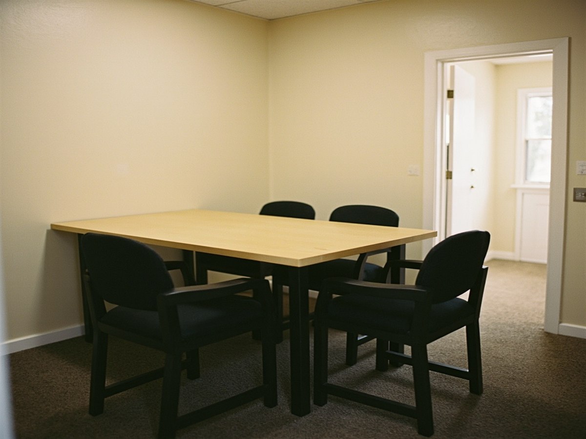 An empty meeting room with a wooden table and three chairs, plain walls and a door standing open onto a corridor.